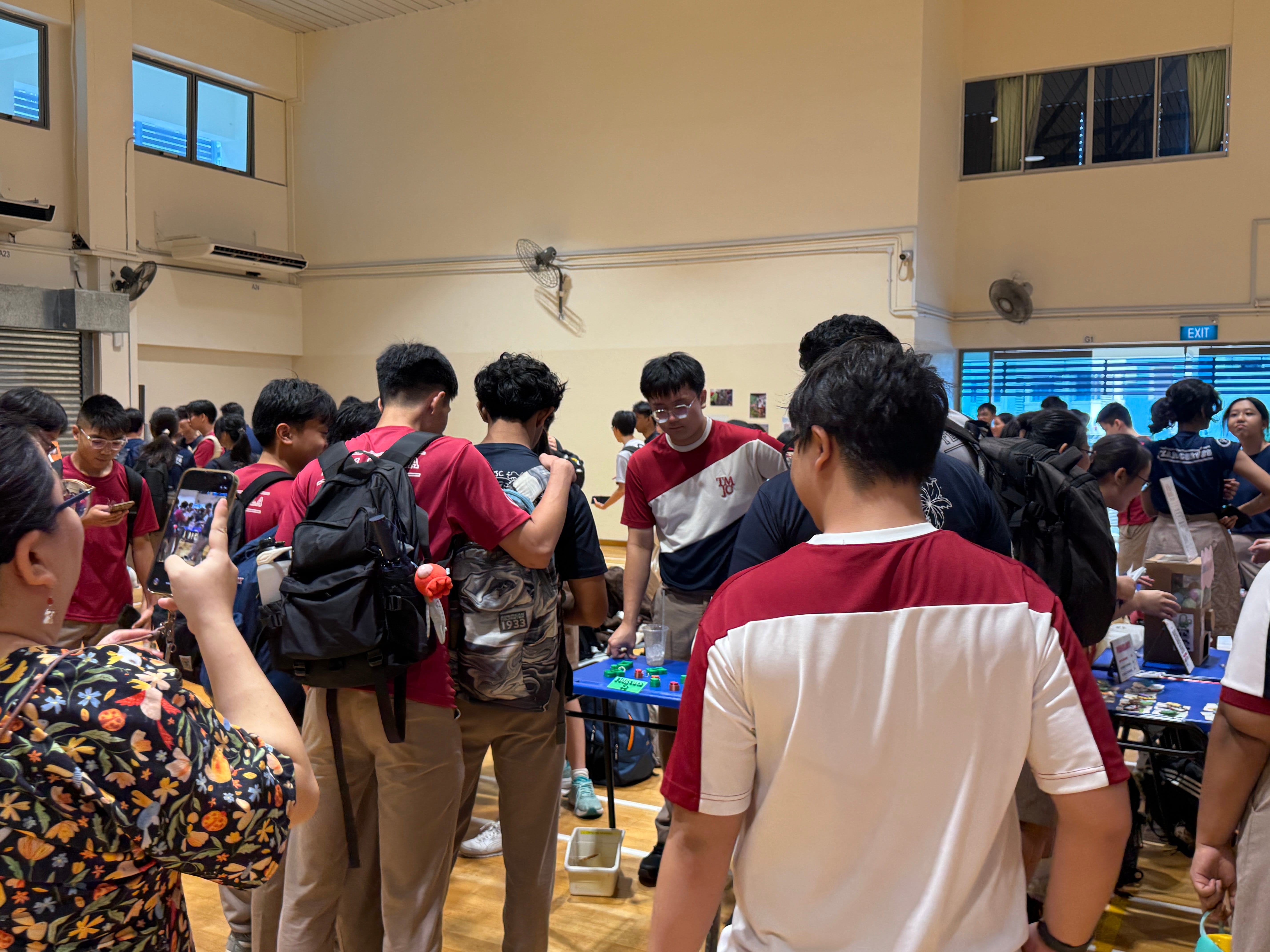 A crowded hall with students browsing a table of handmade items, with one student in a TMJC shirt manning the stall.